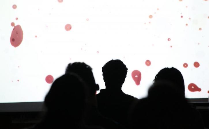 Three silhouettes looking at bright white screen with red dots  